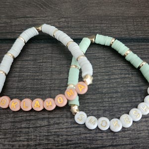 May include: Two beaded bracelets with the words "OKLAHOMA" and "SMOKE SHOW" spelled out in gold letters on white beads. The "OKLAHOMA" bracelet has pink beads and the "SMOKE SHOW" bracelet has light green beads.