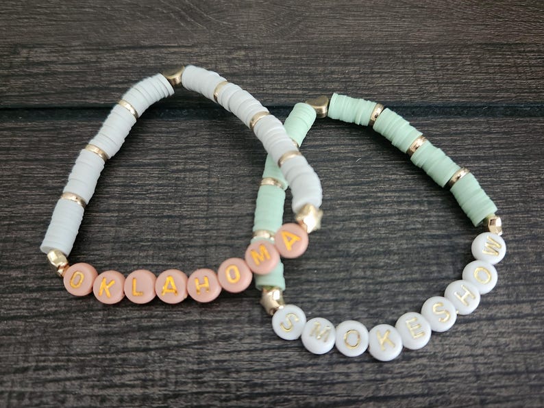 May include: Two beaded bracelets with the words "OKLAHOMA" and "SMOKE SHOW" spelled out in gold letters on white beads. The "OKLAHOMA" bracelet has pink beads and the "SMOKE SHOW" bracelet has light green beads.