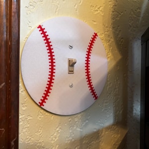 Könnte beinhalten: Eine Lichtschalterabdeckung im Baseball-Design. Die weiße Abdeckung hat rote Nähte, die einem Baseball nachempfunden sind. Sie verfügt über einen Standard-Kippschalter und ist an einer strukturierten Wand montiert.