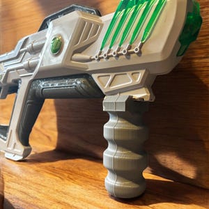 May include: A futuristic toy ray gun in shades of grey and white, with green translucent accents. The toy gun features a detailed design with a ribbed handle and a series of green, blade-like elements along the top.