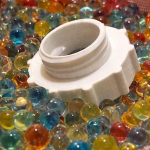 Peut inclure: Un bouchon en plastique blanc, circulaire et fileté, doté d'un bord extérieur cranté, partiellement immergé dans une collection dense de petites perles d'eau translucides et multicolores, jaunes, orange, bleues et transparentes.