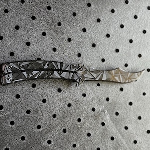 May include: A black butterfly knife with a geometric pattern on the blade. The knife is closed and lying on a black surface with small holes.