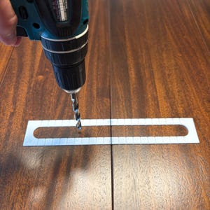 May include: A teal and black cordless power drill positioned over a white plastic drilling template guide with evenly spaced rectangular slots, resting on a dark brown wood surface with a visible vertical grain pattern and a seam.