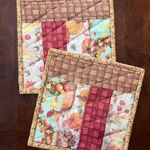 May include: Two square quilted pot holders featuring a patchwork design with autumn-themed fabric. The patterns include pumpkins, fall leaves, acorns, and a brown woven texture. Each piece is finished with a decorative border of small leaves.