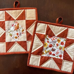 May include: Two square quilted pot holders with a fall theme. The pot holders feature a patchwork design with rust-colored and cream-colored fabrics. One has a squirrel and the other has leaves and flowers.