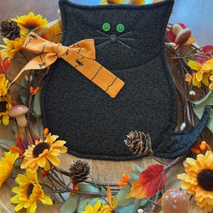 Op de afbeelding: Een zwart katvormig kussen met groene knoopogen en geborduurde snorharen. Het heeft een oranje strik met spinnenafbeeldingen. Het kussen is omgeven door een krans van zonnebloemen, herfstbladeren en dennenappels, wat een Halloween-thema creëert.