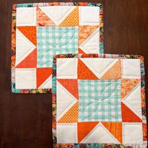 May include: Two square quilted potholders featuring a central star pattern in shades of orange, white, and light teal gingham. The borders are decorated with a colorful print of pumpkins and autumn foliage on a dark background.