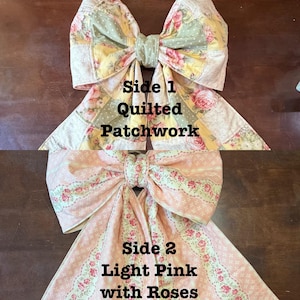May include: Two large fabric bows. Side 1 features a quilted patchwork design with yellow, pink, and green floral patterns. Side 2 is light pink with a rose pattern. The bows are tied in the center with a matching fabric.