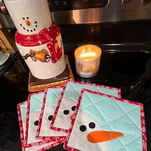 May include: A snowman-themed kitchen set featuring a ceramic cookie jar, a lit candle, and four square quilted pot holders. The pot holders have a snowman face design with a blue hat, black eyes, and an orange carrot nose.