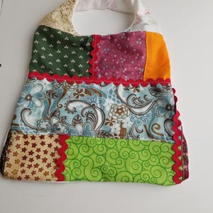 May include: A patchwork baby bib with a variety of fabric patterns and colors. The bib features a floral print, stars, and swirls, with red scalloped trim. The bib has a white terry cloth backing and a neck strap.