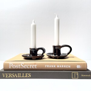 Peut inclure: Deux petits bougeoirs en céramique noire avec poignées, chacun contenant une bougie blanche. Les bougeoirs sont posés sur une pile de livres, dont un intitulé "PostSecret" et un autre "Versailles."