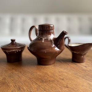 Könnte beinhalten: Ein braunes Keramik-Set aus Teekanne, Milchkännchen und Zuckerdose mit einem Tropf-Glazureffekt. Das Set steht auf einem Holztisch.
