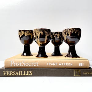 Puede incluir: Cuatro copas de cerámica con base marrón oscuro y un esmalte dorado-marrón goteante. Las copas están dispuestas encima de dos libros, uno titulado "PostSecret" y el otro "Versalles."