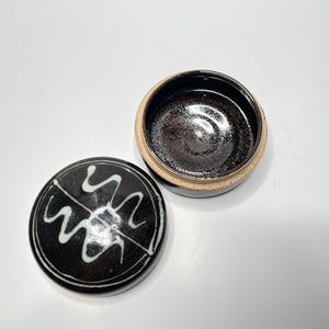 May include: A black ceramic lidded box with a white swirl design on the top. The lid is off and shows the inside of the box, which is also black.