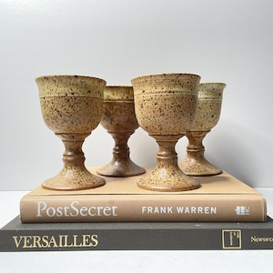Puede incluir: Cuatro copas de cerámica moteada con bases de pedestal. Las copas son una mezcla de marrón y beige, con una superficie texturizada. Están dispuestas encima de dos libros, uno titulado "PostSecret" y el otro "Versailles".