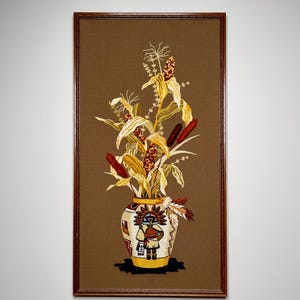 May include: A framed embroidery depicting a vase with a Native American design and a bouquet of dried corn stalks and reeds.