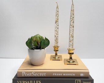 Puede incluir: Dos candelabros dorados con velas transparentes incrustadas con escamas doradas. Las velas están sobre una pila de dos libros, uno beige y otro gris oscuro. Una pequeña planta suculenta verde en maceta está delante de las velas.