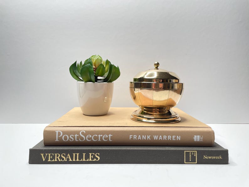 Peut inclure: Un bol d&eacute;coratif en m&eacute;tal dor&eacute; avec un couvercle pos&eacute; sur deux livres. Le livre du dessus est beige et porte l'inscription "PostSecret FRANK WARREN". Le livre du dessous est gris et porte l'inscription "VERSAILLES Newsweek". Une petite plante en pot avec des feuilles vertes est plac&eacute;e &agrave; gauche du bol.