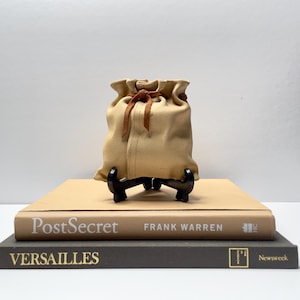 Peut inclure: Un petit sac à cordon beige avec des liens en cuir brun est posé sur un support noir. Le support est placé sur deux livres, l'un beige et l'autre gris. Le livre beige porte le titre "PostSecret" et l'auteur "Frank Warren". Le livre gris porte le titre "VERSAILLES" et le texte "Newsweek" sur le dos.