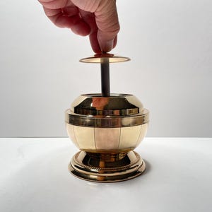 Peut inclure: Un cendrier en m&eacute;tal dor&eacute; avec un corps rond en forme de globe et un couvercle amovible. Le couvercle est soulev&eacute; par une main.