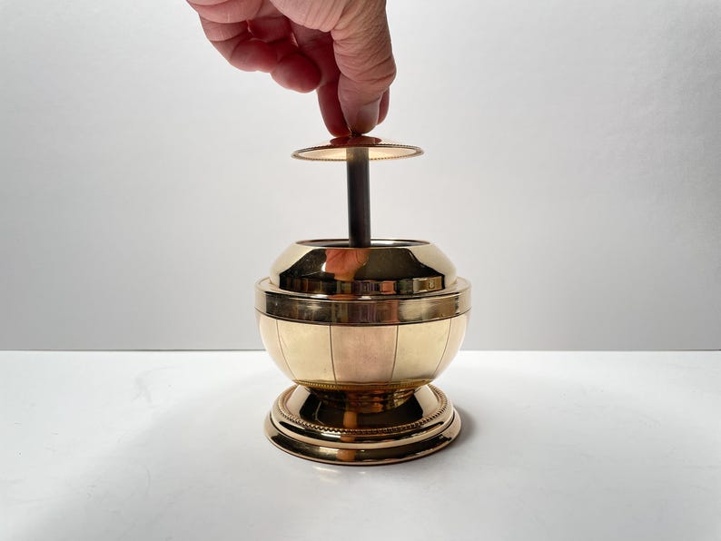 Peut inclure: Un cendrier en m&eacute;tal dor&eacute; avec un corps rond en forme de globe et un couvercle amovible. Le couvercle est soulev&eacute; par une main.