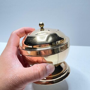 Peut inclure: Un r&eacute;cipient en m&eacute;tal dor&eacute; avec un couvercle bomb&eacute; et un petit bouton sur le dessus. Le r&eacute;cipient a une bande d&eacute;corative autour du milieu et une base avec un bord perl&eacute;.