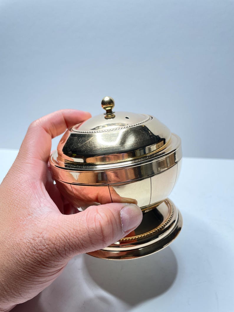 Peut inclure: Un r&eacute;cipient en m&eacute;tal dor&eacute; avec un couvercle bomb&eacute; et un petit bouton sur le dessus. Le r&eacute;cipient a une bande d&eacute;corative autour du milieu et une base avec un bord perl&eacute;.