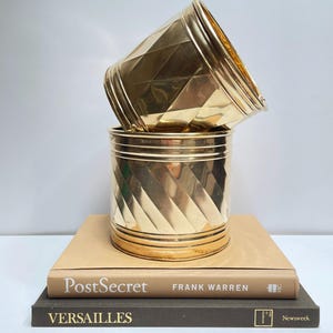 May include: Two gold metal planters with a diamond pattern stacked on top of each other. The planters are sitting on top of two books, one with the title "PostSecret" and the other with the title "VERSAILLES".