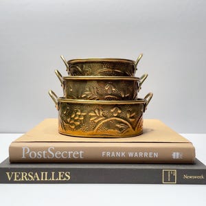 Puede incluir: Tres cuencos de latón con diseños florales apilados uno encima del otro. Los cuencos están sobre dos libros, uno beige y otro gris. El libro beige tiene el título "PostSecret" y el libro gris tiene el título "Versailles".