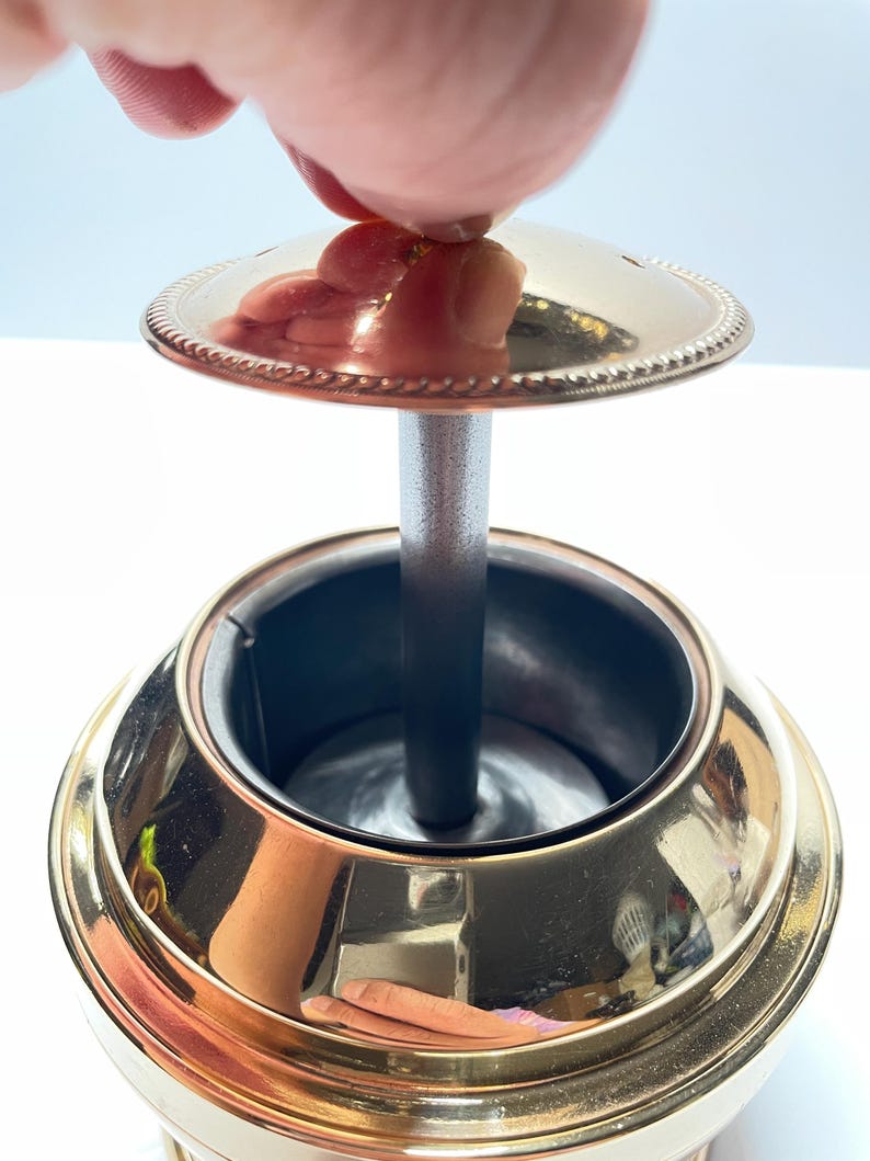 Peut inclure: Un cendrier en m&eacute;tal dor&eacute; avec un couvercle amovible. Le couvercle est orn&eacute; d'un motif de corde d&eacute;coratif autour du bord. Le cendrier a un cylindre central pour contenir les cigarettes.
