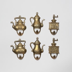 May include: Six gold-toned metal hooks shaped like coffee pots and teapots. The hooks are decorative and have a vintage style.