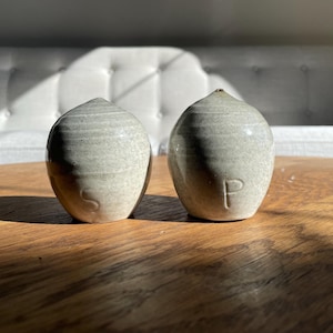May include: A set of two ceramic salt and pepper shakers in a light grey colour with a speckled finish. The shakers are round and have a slightly textured surface. The word "S" is visible on one shaker and the word "P" is visible on the other shaker.