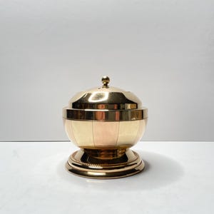 Peut inclure: Un r&eacute;cipient en m&eacute;tal dor&eacute; avec un couvercle et une base ronde. Le r&eacute;cipient pr&eacute;sente un motif d&eacute;coratif de lignes verticales.