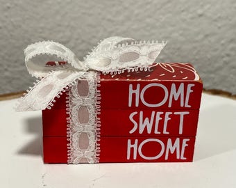 Op de afbeelding: Drie rode houten blokken gestapeld op elkaar. Elk blok heeft witte letters. Het bovenste blok zegt "HOME", het middelste blok zegt "SWEET" en het onderste blok zegt "HOME". De blokken zijn aan elkaar gebonden met een wit kantlint.