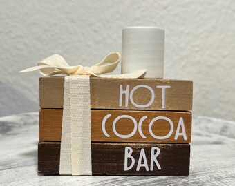 Könnte beinhalten: Ein Holzschild mit den Worten "Hot Cocoa Bar" übereinander gestapelt. Das Schild ist mit einem weißen Band und einem weißen Marshmallow verziert.