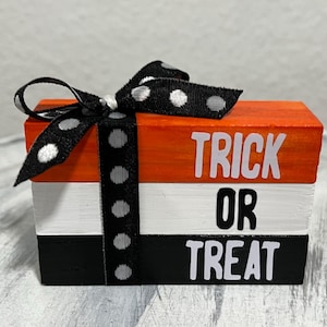 May include: A Halloween-themed wooden block decoration. The stacked blocks are painted orange, white, and black, with the words "TRICK OR TREAT" in white. A black ribbon with white polka dots is tied around the blocks.