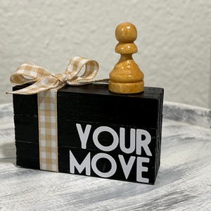 May include: A black rectangular wooden block with the white text YOUR MOVE printed on the front. A light tan and white gingham ribbon is tied around the block in a bow. A single light brown wooden chess pawn sits on top of the black block.