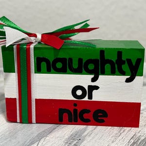 May include: A rectangular block painted in Christmas colors: green, white, and red. The block has the words "naughty or nice" in black. A decorative bow with red, white, and green ribbons sits on top.