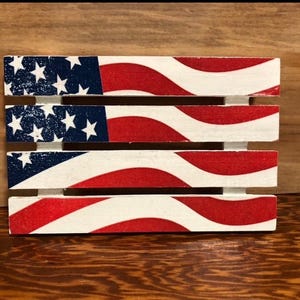 May include: A wooden pallet painted with an American flag design. The pallet is made of four horizontal slats with red, white, and blue stripes. The stars are white on a blue background.