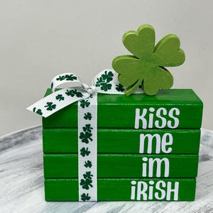 May include: A green wooden block stack with the text "Kiss me im iRISH" in white. A white ribbon with green shamrocks is tied around the blocks. A green wooden shamrock sits on top.