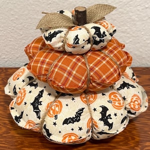 May include: A decorative, fabric pumpkin in a tiered design, perfect for Halloween. The pumpkin features orange plaid and cream-colored fabric with black bat and jack-o'-lantern prints. It is adorned with a wooden stem and burlap leaves, creating a festive autumn decoration.