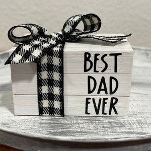 May include: A white wooden block sign with the words "Best Dad Ever" stacked on top of each other. The sign is decorated with a black and white checkered ribbon.