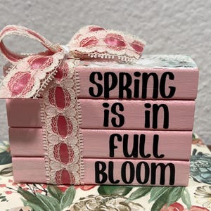 May include: A stack of four pink wooden blocks with the words "SPRING is in FULL BLOOM" in black lettering. A pink and white lace ribbon and bow adorn the top. The blocks sit on a floral patterned surface.