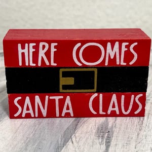 May include: A red wooden block sign with the words "HERE COMES SANTA CLAUS" in white lettering. The sign has a black belt with a gold buckle design in the center, creating a festive holiday decoration.