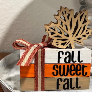 May include: A decorative wooden block set with the words "Fall Sweet Fall" in black lettering. The blocks are painted white, orange, and tan, and are tied with a striped ribbon. A wooden leaf cutout sits on top.