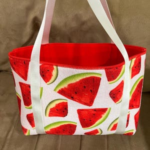 May include: A tote bag featuring a repeating watermelon slice pattern on a white background. The bag has a bright red interior lining and two white fabric handles attached to the exterior. The watermelon slices are red with green rinds.