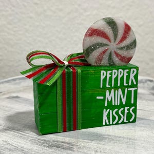 May include: A green wooden block with the words "PEPPER -MINT KISSES" in white. A red and green striped ribbon is tied around the block. A peppermint candy with red, white, and green stripes sits on top.
