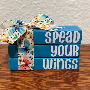 May include: A stack of three teal wooden blocks with the words "SPEAD YOUR WINGS" in white. A colorful butterfly ribbon is tied around the blocks, adding a decorative touch. The blocks are on a wooden surface.