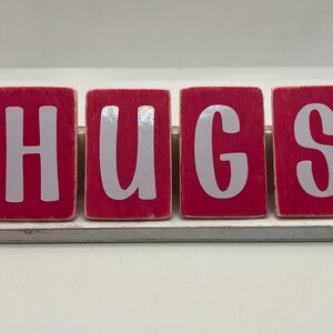 May include: A decorative wooden sign with the word "HUGS" spelled out in large, white letters. The letters are on individual red blocks, arranged on a white and gray wooden base. The sign is a charming home decor item.