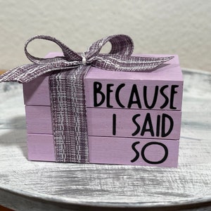 May include: A stack of three small wooden blocks painted lavender with black lettering. The blocks are tied together with a purple and white ribbon. The text on the blocks reads "Because I Said So".